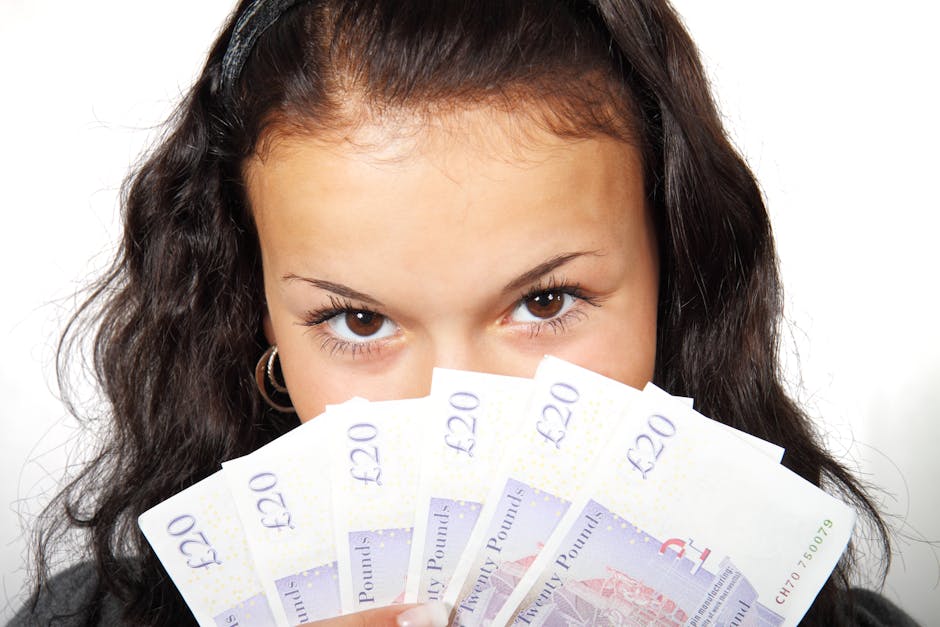 A young woman with long, dark, curly hair and light brown eyes is holding multiple £20 banknotes in front of her face, partially covering her mouth and nose. She is looking directly at the camera with a neutral expression. The background is plain white, and she appears to be indoors. The image does not depict house removals or moving services, but could be used in contexts related to costs, payments, or finance in a home relocation setting.