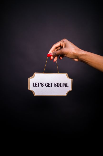 A person's hand with neatly manicured nails painted red is holding a small, rectangular, beige sign with a thin golden string. The sign displays the message 'LET'S GET SOCIAL' in bold black capital letters. The background is solid black, providing high contrast and focus on the hand and sign. The image emphasizes a social media or networking theme, with the hand positioned centrally and the sign hanging below, creating a balanced composition. This visual can be associated with promoting connection and engagement, relevant to house removals and moving services by highlighting communication and customer interaction. The lighting is even, with no visible shadows, ensuring clarity of the sign and hand.
