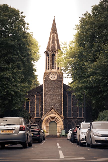 A narrow street leading directly to a traditional stone church with a tall, pointed spire and a clock face just below the spire. The church is framed by mature trees on either side, creating a symmetrical appearance, and the sky appears overcast. In the foreground, a line of parked cars is visible along both sides of the street. The street is part of a home relocation process, with the scene suggesting a quiet residential area. The image supports the context of house removals and moving logistics, as part of a service offered by Man with Van Pinner, with the church in the background providing a local landmark near Pinner. The perspective emphasizes the process of loading furniture and boxes into a vehicle, with the scene capturing the typical environment for a house move within the area linked to Harrow Council permits.