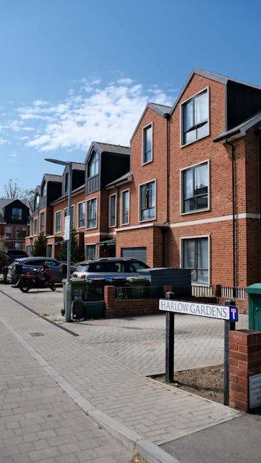 An outdoor view of a residential street in Harlow Gardens featuring a row of modern multi-storey brick apartment buildings with dark-colored window frames and sloped roofs, under a clear blue sky with some scattered clouds. In the foreground, a paved sidewalk runs alongside the street, with a small signpost indicating Harlow Gardens and a blue road sign. Several parked cars are positioned along the curb, including a black hatchback and a grey sedan. Near the street, there are green waste bins and a black wheelie bin, suggesting waste collection areas. This scene may be relevant for house removals or relocation services, such as those offered by Man with Van Pinner, especially when planning furniture transport or home relocation logistics involving apartment buildings and street parking.