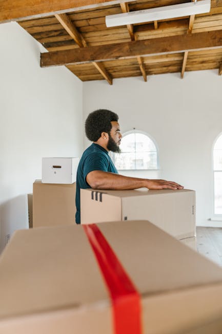 A man with dark curly hair and a beard, wearing a blue T-shirt, stands inside a spacious, well-lit room with a high, exposed wooden ceiling and white walls. He is positioned behind several large cardboard moving boxes, one with red packing tape, which are stacked in front of him and appear to be prepared for a home relocation. The room features two large arched windows allowing natural light to enter, and the floor is wooden. In the background, a partially visible white wall and the structure of the ceiling are apparent. The man looks focused while engaged in the loading process, representing a typical furniture transport and packing stage of a house removal, with the boxes ready to be loaded onto a van. This scene is associated with the services provided by Man with Van Pinner, a company specialising in removals and house moving services, highlighting the packing and logistics aspect of local house removals in Pinner. The environment suggests an organized, professional approach to home relocation, with attention to careful handling of packing materials during the furniture transport process.
