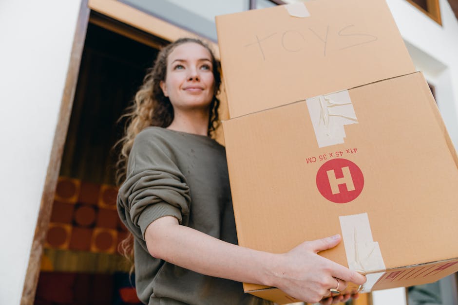 A young woman with long curly hair and wearing a grey sweatshirt is inside a room, holding a cardboard box labeled 'toys' and another larger box with red and white markings, part of her home relocation or packing process. The boxes are made of brown cardboard, reinforced with clear packing tape. She stands near a doorway or entrance, with a wooden frame visible in the background, and an interior wall with red circular patterns. Natural light illuminates her face, and she appears to be preparing or organizing items for moving as part of a house removal carried out by Man with Van Pinner, a professional removals service. The scene depicts the loading or packing stage of furniture transport, with focus on packaging materials, the boxes' positioning, and the indoor environment that supports the moving logistics related to the 'Moving Out of Pinner Village Estate' checklist.