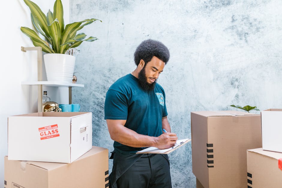 A man with dark curly hair and a beard, wearing a blue T-shirt, stands inside a spacious, well-lit room with a high, exposed wooden ceiling and white walls. He is positioned behind several large cardboard moving boxes, one with red packing tape, which are stacked in front of him and appear to be prepared for a home relocation. The room features two large arched windows allowing natural light to enter, and the floor is wooden. In the background, a partially visible white wall and the structure of the ceiling are apparent. The man looks focused while engaged in the loading process, representing a typical furniture transport and packing stage of a house removal, with the boxes ready to be loaded onto a van. This scene is associated with the services provided by Man with Van Pinner, a company specialising in removals and house moving services, highlighting the packing and logistics aspect of local house removals in Pinner. The environment suggests an organized, professional approach to home relocation, with attention to careful handling of packing materials during the furniture transport process.