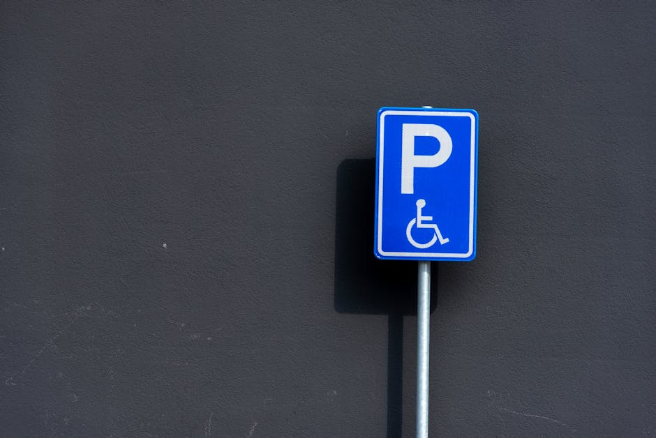A blue parking sign with a white 'P' and a wheelchair symbol indicating accessible parking, mounted on a silver metal pole against a dark grey wall. The shadow of the sign is cast onto the wall. This scene is outdoors, likely on a pavement or driveway adjacent to a building, and is relevant to parking arrangements for home relocation or furniture transport in the context of house removals. The sign is positioned slightly to the right of the image, with part of the pavement visible at the bottom. The lighting is natural, illuminating the sign clearly for accessibility purposes, supporting the process of loading or unloading furniture during a move with professional removals. Occasionally, Man with Van Pinner advising on parking permits for Pinner moves can benefit from clear signage like this for access during classified parking suspension applications.