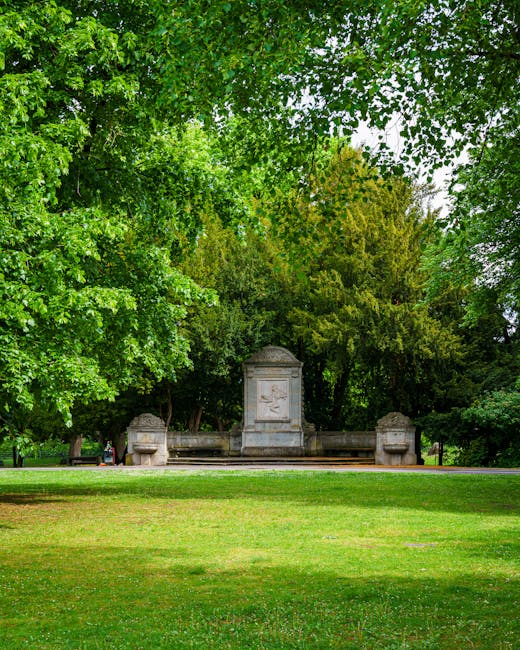 A view of a park area with lush green trees providing ample shade, framing a stone monument or memorial in the background. The monument is flanked by two large stone planters or decorative features, and there are benches visible near the monument, suggesting an area for visitors to sit and relax. The foreground consists of well-maintained grass leading up to the monument, with a pathway visible at the edge of the image. The scene is well-lit with natural daylight, indicating a clear or partly cloudy day. The image illustrates the peaceful environment of Pinner Memorial Park, which is a suitable location for house removals or relocation services, and the scene is captured by Man with Van Pinner, a professional moving company that offers removals and moving logistics in the area.