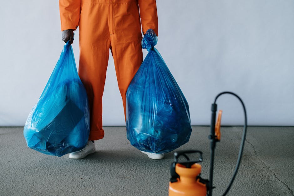 A person dressed in orange work overalls is standing indoors on a grey carpeted floor, holding two large blue plastic rubbish bags filled with discarded items, likely as part of a home clearance or packing process for a house move. The individual's lower body is visible, with white shoes and their hands gripping the top handles of each bag. In the foreground, there is a black and orange sprayer or pest control device connected to an extension cord, indicating cleaning or disposal equipment used during the packing or moving procedure. The background features a plain, light-colored wall, suggesting a neutral environment such as a garage or utility room, with natural or indoor lighting illuminating the scene. This image reflects elements involved in packing, waste disposal, or pre-move preparations as part of a professional house removal service by Man with Van Pinner, emphasizing proper handling and transport of waste materials in the context of relocating.