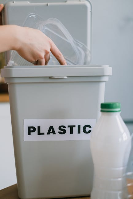 A close-up image shows a person's hand lifting the lid of a grey plastic recycling bin labeled 'PLASTIC'. Inside the bin, transparent plastic packaging is visible. Next to the bin, there is a partially visible white plastic bottle with a green cap. The scene is set indoors, possibly in a kitchen or utility area, with a neutral background. This image relates to the sorting and disposal of recyclable materials as part of home relocation or moving preparations. Man with Van Pinner offers removals services that include waste disposal and proper recycling procedures, important aspects when planning a house move to comply with local rules and avoid fines. The overall setting suggests the process of packing, organizing, and responsibly managing waste during a moving project.