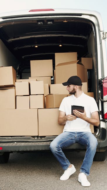 A man wearing a black cap, white t-shirt, and blue jeans is seated on the pavement beside the open rear of a van loaded with various packed cardboard boxes in different sizes. The boxes are stacked ne