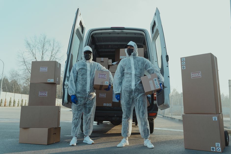 A man wearing a black cap, white t-shirt, and blue jeans is seated on the pavement beside the open rear of a van loaded with various packed cardboard boxes in different sizes. The boxes are stacked ne