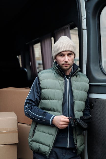 A man wearing a white t-shirt, blue jeans, white sneakers, and a black cap is walking beside a white cargo van with its sliding door open on a suburban street. He is carrying three stacked cardboard b