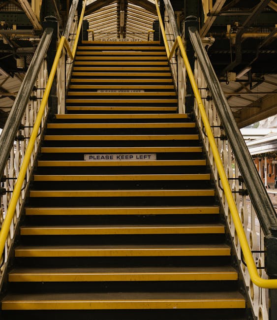 A set of indoor industrial stairs with yellow handrails and black and yellow anti-slip treads, leading upwards in a warehouse or loading area. The stairs are constructed from metal and are surrounded 