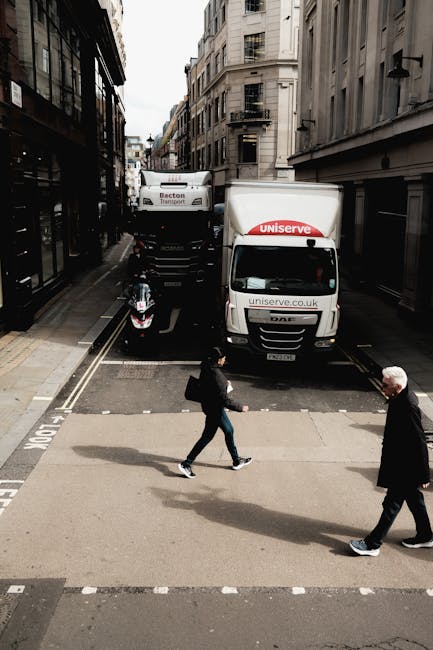 A street scene in an urban area with two large white moving trucks parked parallel to each other on a narrow road, one branded with 'Uniserve' and the other with 'Becton Transport.' In front of the trucks, a black motorcycle is parked on the pavement. Two pedestrians are crossing the road: a woman carrying a shoulder bag and wearing a black coat and jeans, and an older man in a dark jacket and trousers. The surroundings include multi-storey buildings with multiple windows and fire escapes, typical of a city centre environment. The scene appears to capture the loading or unloading process during a home relocation or furniture transport, with the trucks positioned close to the building entrance, indicating a planned move coordinated by [COMPANY_NAME], specialists in removals and moving services in Pinner.
